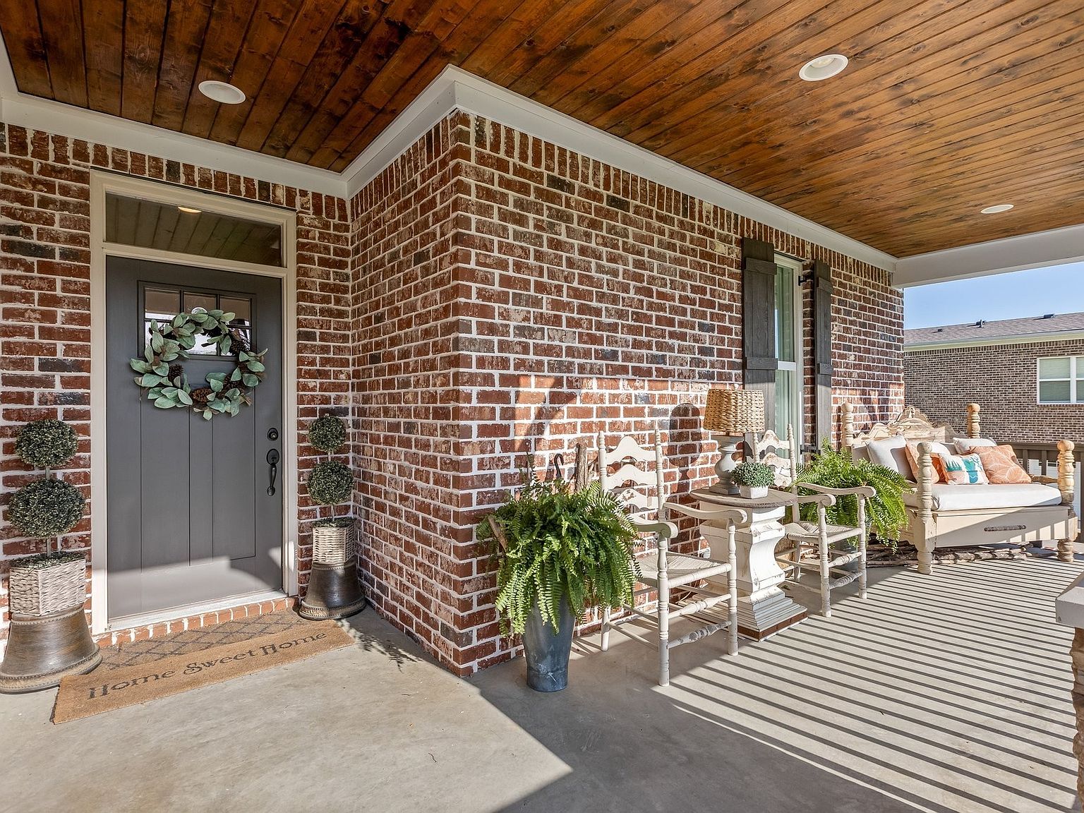 Close view of the front door at 1070 Hideaway Road, a gray panel door with seasonal wreath set into a brick wall, with a Home Sweet Home mat at the threshold and wicker seating to the side